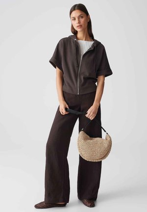 Frau trägt einen schwarzen Kapuzenpullover mit kurzen Ärmeln und Reißverschluss, weite schwarze Hosen, hält eine gewebte, halbmondförmige beigen Handtasche.