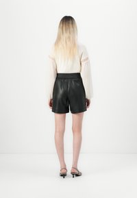 Person med långt blont hår står vänd mot en vit vägg, klädd i en genomskinlig vit långärmad blus, svarta lädershorts och låga klackskor.