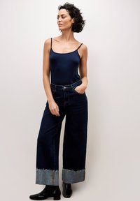 Marineblaues Top ohne Ärmel kombiniert mit weit geschnittenen, dunklen Jeans mit einem ausgefransten Denim-Saum. Schwarze Stiefeletten runden den Look ab.