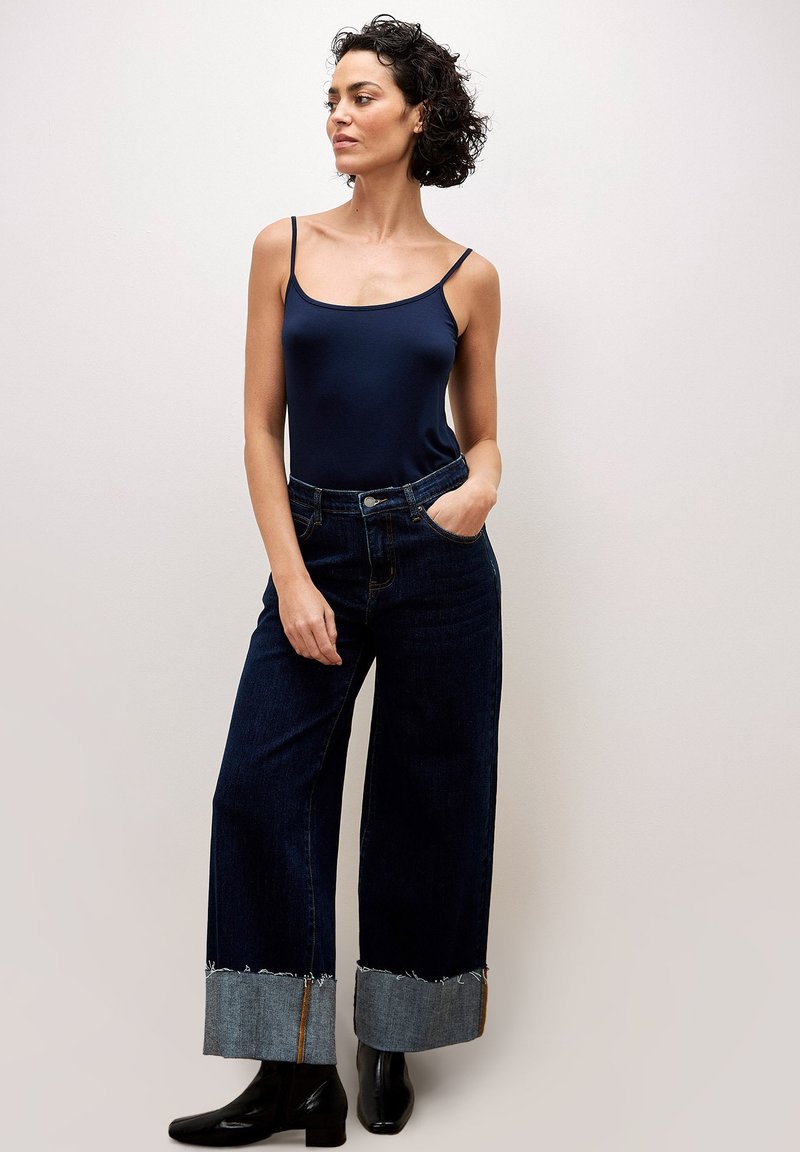 Marineblaues Top ohne Ärmel kombiniert mit weit geschnittenen, dunklen Jeans mit einem ausgefransten Denim-Saum. Schwarze Stiefeletten runden den Look ab.
