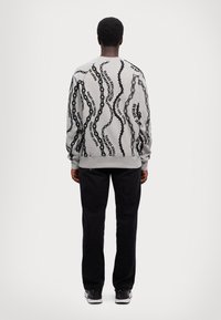 Sudadera gris con un patrón de cadena ondulada negra, con puños y cinturilla acanalados, combinada con pantalones negros y zapatillas, vista desde atrás.