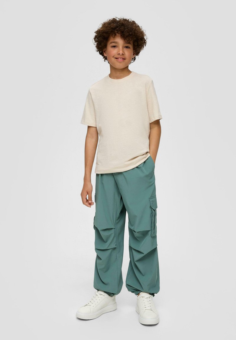 Beige Kurzarm-T-Shirt kombiniert mit türkisfarbenen Cargo-Hosen mit Seitentaschen, gerafften Bündchen und weißen Sneakern. Neutraler Hintergrund.