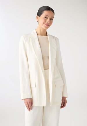 Donna che indossa un blazer crema strutturato e pantaloni coordinati sopra una maglia beige lavorata a maglia, in piedi davanti a uno sfondo bianco semplice.