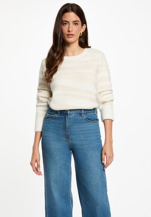 Maglione crema con strisce beige chiaro, scollatura rotonda e polsini a coste, abbinato a jeans blu a vita alta e gamba larga. Texture morbida.