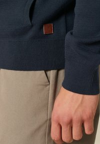 Marineblauer Strickpullover mit gerippten Bündchen und Saum, ausgestattet mit einem Logo-Patch aus braunem Leder. Kombiniert mit hellbeigen Hosen, die über Taschendetails verfügen.