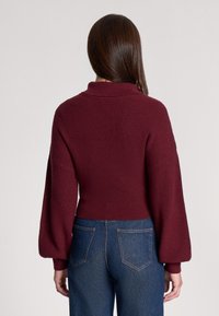 Burgundowy sweter z dzianiny o wysokim kołnierzu, balonowych rękawach i żebrowanej fakturze, w zestawieniu z ciemnoniebieskimi dżinsami z wysokim stanem.