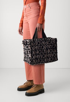 Personne portant un pantalon rose, des bottes montantes camel et un haut orange à manches longues, tenant un sac fourre-tout noir et beige avec un texte à motifs.