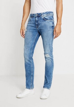 Mann trägt slim-fit hellblaue Denim-Jeans mit kleinen Rissen am Oberschenkel und Knie, kombiniert mit weißen Sneakers und weißem T-Shirt.