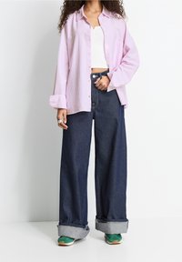 Camicia button-up a righe verticali rosa, canotta bianca aderente, jeans scuri a gamba larga con orli arrotolati e sneakers verdi.