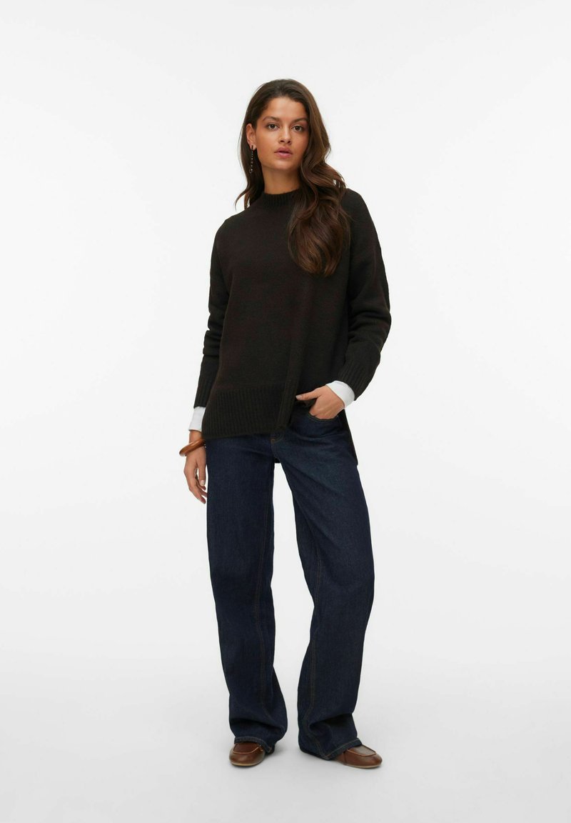 Mørkebrun sweater med ribbet kant og lyse manchetter, parret med brede mørke denimjeans og brune sko.