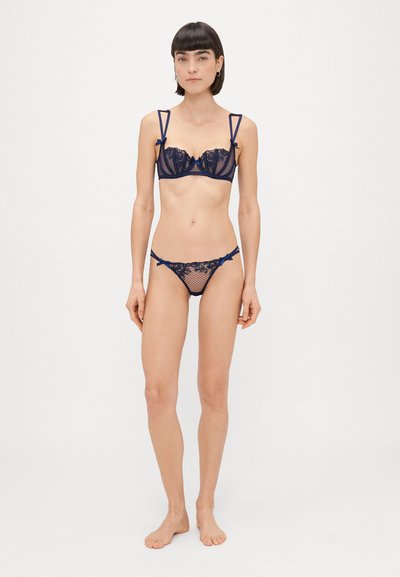 Jūras lašu mežģīņu komplekts, kas ietver viegli polsterētu krūšturi ar izgriezumiem un lentītēm, kopā ar atbilstošām bikī apakšdaļām, rotātām ar mežģīņu detaļām.