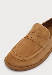 Mocassin en daim beige avec coutures décoratives et détails perforés, bout arrondi et semelle en caoutchouc beige sur fond blanc.