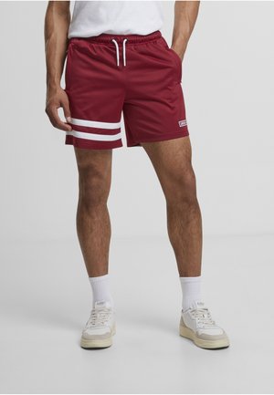 Uomo che indossa pantaloncini sportivi color bordeaux con due strisce bianche sulla gamba sinistra, calzini bianchi e scarpe da ginnastica bianche, in piedi contro uno sfondo semplice.
