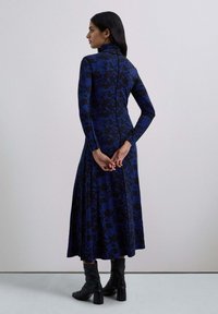 Abito a maniche lunghe di color blu scuro con motivo astratto nero. Corpino aderente che si trasforma in una gonna fluida. Indossato con stivali neri al ginocchio.