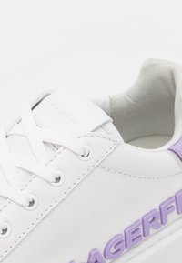 Baskets blanches avec lacets blancs et lettrage violet sur le côté, col rembourré et détails cousus sur la tige.