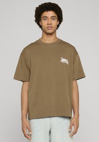 Brązowy, oversize'owy bawełniany t-shirt z krótkimi rękawami, z białym nadrukiem graficznym na lewej piersi. W zestawie z jasnoniebieskimi szortami jeansowymi.