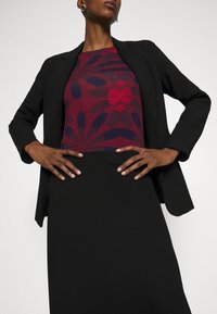 Žena v černém blejzru a sukni, s červeným a námořnickým květinovým vzorem na topu, stojí s rukama na bok proti jednobarevnému pozadí.