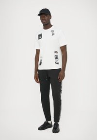 White graphic t-shirt with various black patches, paired with black sweatpants featuring side stripes and black sneakers.