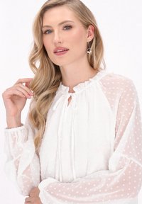 Weiße, gepunktete Bluse mit einem Rüschenausschnitt und Kordelzug. Lange, durchscheinende Ärmel, die sich an den Handgelenken sammeln. Voluminös mit einer leichten Textur.