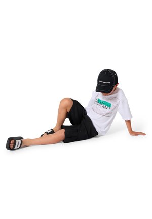 T-shirt blanc avec imprimé graphique, short cargo noir, sandales noires avec logo blanc, position assise, portant une casquette noire avec logo.