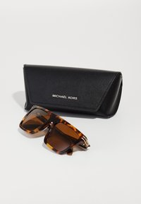 Tortoiseshell sončna očala s svetlo rjavimi lečami in zlatim okovjem, postavljena ob črni usnjeni etui z embossiranim napisom "MICHAEL KORS."