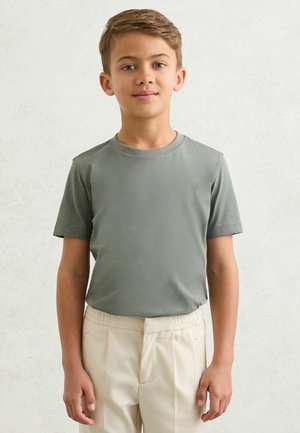 Rövid barna hajú fiú szürke pólót és bézs nadrágot visel, sima világos háttér előtt áll.