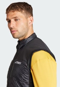 Ärmelleste schwarze isolierte Weste mit gestepptem Design, versehen mit dem "TERREX"-Logo. Getragen über einem langärmeligen gelben Hemd.