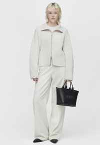 Giacca zip-up ribbed color beige chiaro con un ampio colletto, abbinata a pantaloni a gamba larga color crema e una piccola borsa nera con testo in rilievo.