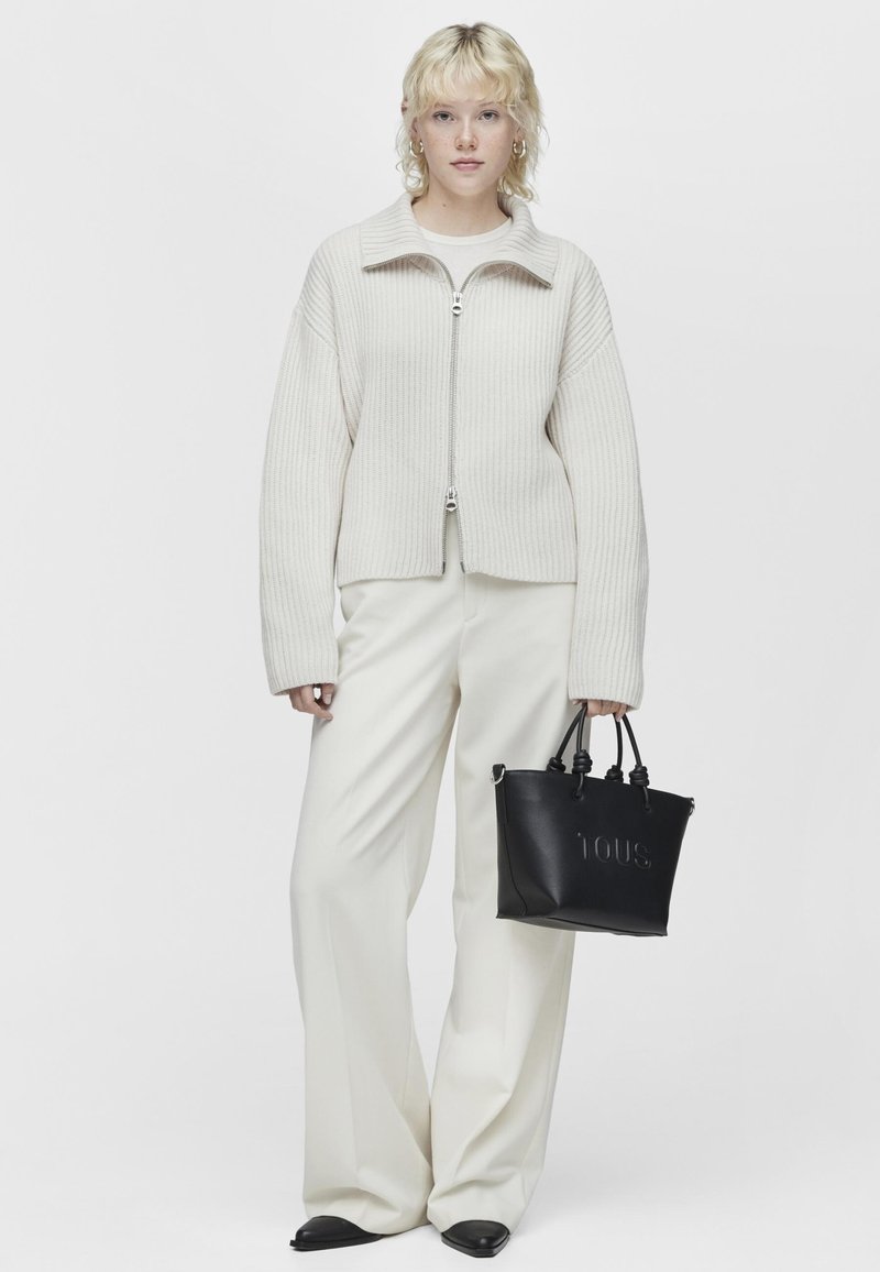Giacca zip-up ribbed color beige chiaro con un ampio colletto, abbinata a pantaloni a gamba larga color crema e una piccola borsa nera con testo in rilievo.