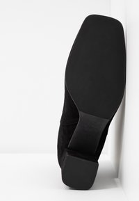 Botte en daim noir positionnée avec la semelle vers le haut contre une surface blanche, montrant un talon carré et une semelle extérieure lisse et plate avec un texte embossé.