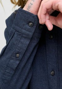 Person wearing a dark blue buttoned shirt with a visible tattoo on wrist partially covered by the shirt cuff.