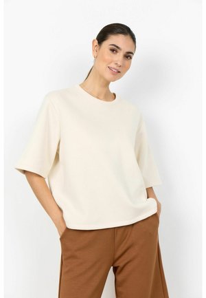 Ženska s temnimi lasmi v bež preveliki kratkoročni majici in rjavih hlačah, z rokami v žepih, stoji pred belim ozadjem.