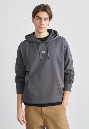 Felpa con cappuccio grigia in cotone con tasca a marsupio, cappuccio con coulisse, polsini a coste e piccolo logo bianco sul petto. Indossata sopra una maglietta nera.