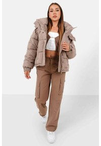 Bruine gewatteerde jas met een hoge kraag, gecombineerd met beige cargo broek met zijzakken. Witte sneakers maken de outfit compleet.