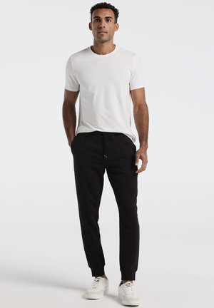 Witte t-shirt met korte mouwen gecombineerd met zwarte joggerbroeken en witte sneakers. De joggers hebben een elastische tailleband en manchetten bij de enkels.