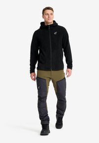 Schwarze Fleece-Softshell-Jacke mit seitlichen Taschen, kombiniert mit grünen und grauen Outdoor-Hosen mit Knieverstärkungen und Logo-Details. Schwarze Stiefel.