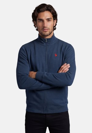 Man with folded arms wearing navy blue zip-up jacket with red logo on chest, standing against plain light background.