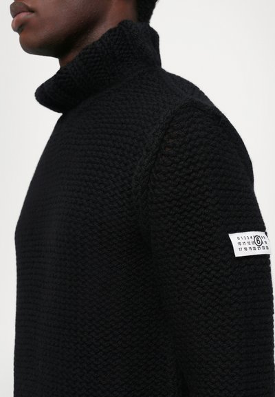 Svart polotröja gjord av texturerad stickad tyg, med hög krage och en lapp med siffror på vänster ärm.