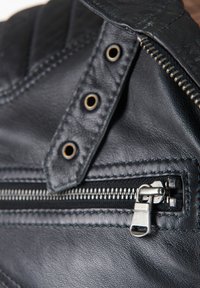 Sort lædermateriale med synlige stikninger, der har en lynlåslomme og dekorative hardwaredetaljer. Nærbillede af den teksturerede overflade.
