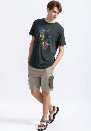T-shirt di cotone verde scuro con stampa grafica colorata, abbinata a pantaloni cargo beige con tasche laterali verdi e sandali neri.