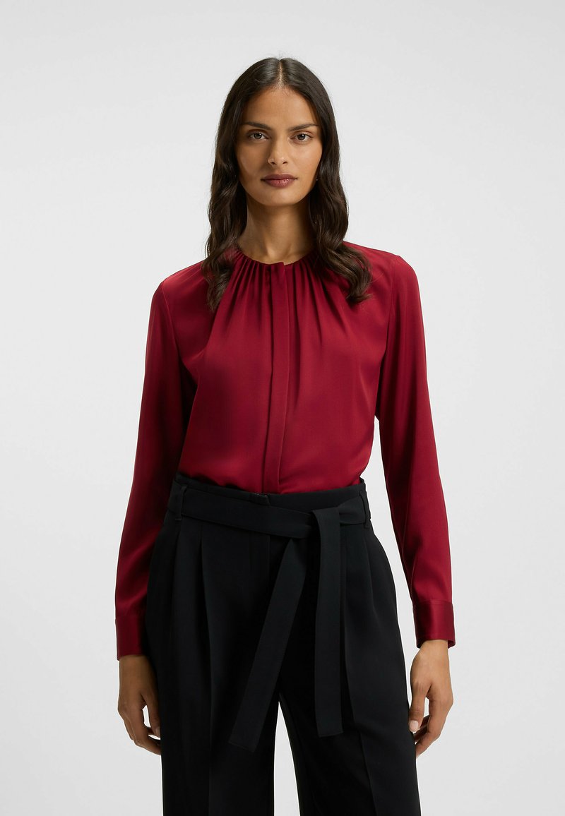 Blusa bordeaux con scollatura rotonda plissettata e maniche lunghe, abbinata a pantaloni neri a vita alta con cintura legata. Tessuto liscio e lucido.