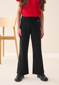 Juodos, platėjančios kelnės su lygiu audiniu ir elastinga juosmens juosta, ant priekio su mažu logotipu. Derinamos su blizgančiais juodais batais.