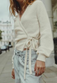 Cream-colored wrap sweater with long sleeves, tied at the waist, paired with light blue denim jeans featuring decorative pocket accents.