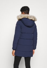 Marinblå puffjacka med strukturerad tyg, foder i päls på huvan och horisontellt kviltat mönster, samt logotypdetalj på ärmen.