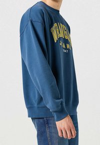 Sweat-shirt bleu coupe décontractée, avec un col rond et des manches longues. Texte graphique jaune à l'avant, avec poignets et ourlet côtelés.