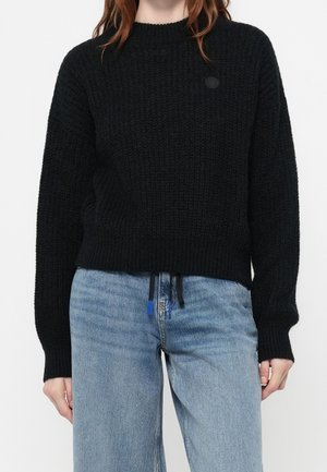 Personne portant un pull noir en tricot à manches longues et un jean bleu clair taille haute avec des détails à cordon.
