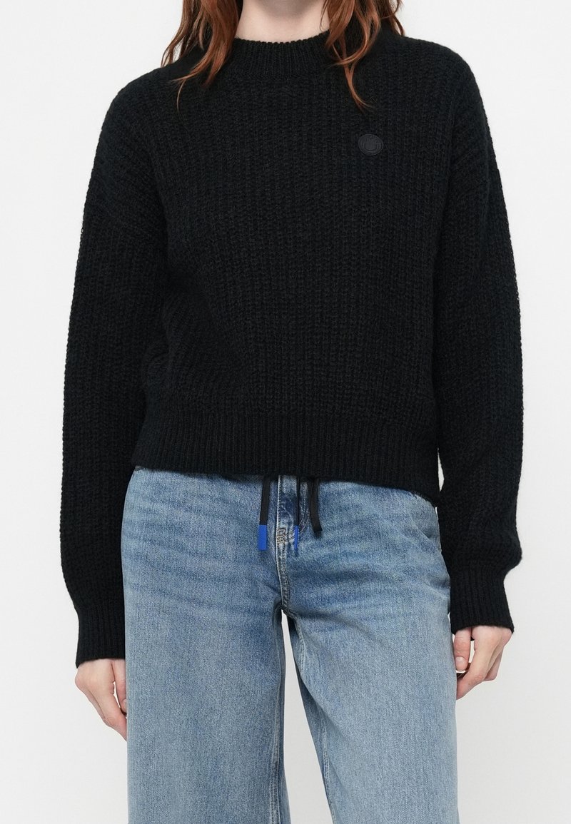 Personne portant un pull noir en tricot à manches longues et un jean bleu clair taille haute avec des détails à cordon.