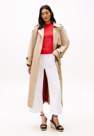 Trench coat beige con interno rosso, indossato sopra un maglione rosso a coste e pantaloni bianchi a gamba larga. Sandali con zeppa neri a cinturini completano il look.
