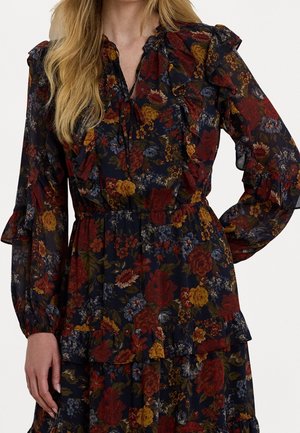 Kvinde iført en mørk blomsterprintet langærmet kjole med flæser, med røde, gule og blå blomster og elastisk talje.