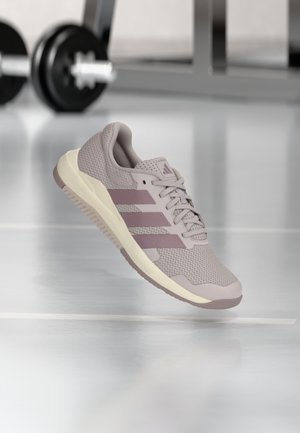 Chaussure de course Adidas violet clair avec tige en mesh et trois bandes, affichée en suspension dans un cadre de salle de sport avec du matériel de fitness en arrière-plan.
